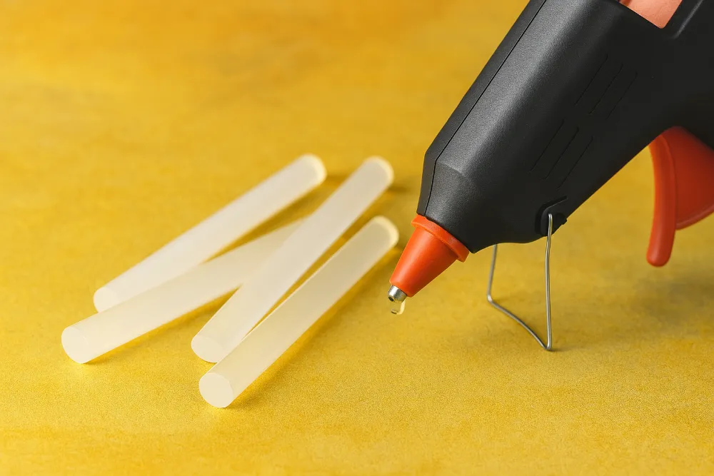 Чорний клейовий пістолет з краплею клею та стрижнями на жовтому фоні