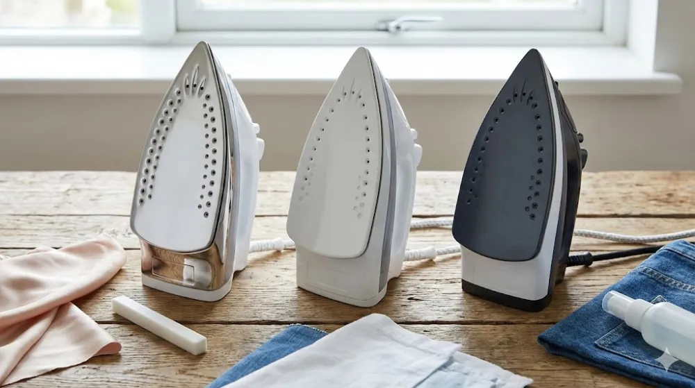 Порівняння підошв прасок: сталь, кераміка, тефлон Три праски з підошвами з нержавійки, кераміки та тефлону на дерев'яному столі зі зразками тканин і засобами догляду